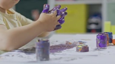Evdeki dağınık ama eğlenceli bir sanat seansı sırasında mor boyayı bütün ellerine sürten küçük bir çocuk.