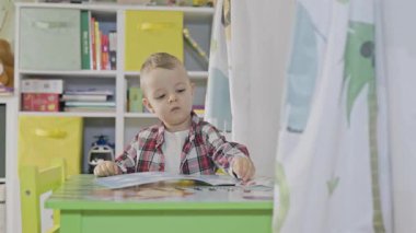 Genç bir çocuk, erken eğitim için renkli oyun odasında yeşil bir masada bir çıkartma kitabıyla oynuyor.