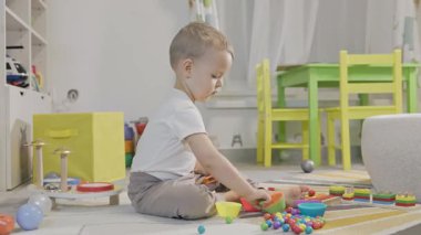 Küçük çocuk yerde oturmuş renkli toplar ve fincanlarla oynuyor, motor becerilerini geliştiriyor.