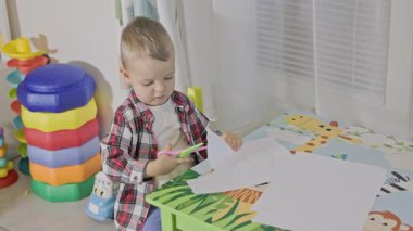 Oyun odasında makasıyla kağıttan bir şekil keserken konsantre olan sevimli küçük çocuk.