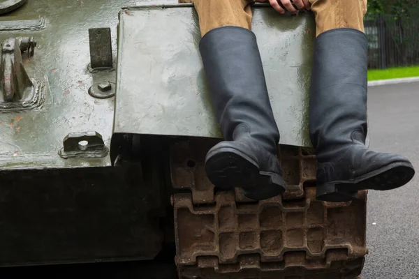 Fragment of the front of an old military tank. A track, an armor plate and the legs of a man in soldiers\' boots are visible. Background.