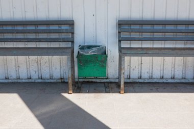 Otobüs durağının yanındaki binanın bir parçası. Ahşap koltuklu metal banklar ve içinde çöp torbası olan yeşil bir çöp kutusu var. Sütun ve metal duvarın gölgesi görünür..