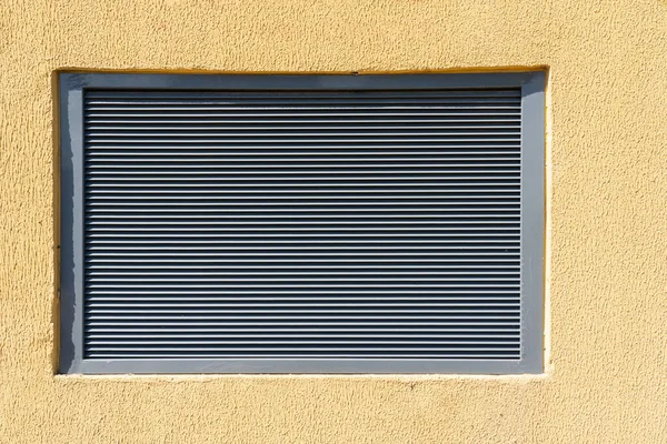 A gray metal window in a yellow stone wall. There is a louvred ventilation grill. Background. Texture.