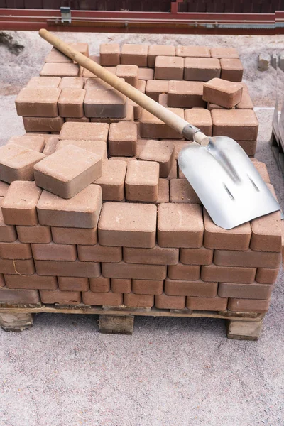 The ocher-colored paving slabs are stacked in neat rows on a wooden pallet. On top is a shovel with a wooden handle. Hepla is covered with a sandy mixture.