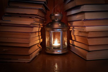 A candle is burning in a vintage metal lantern with glass. There are stacks of old books on either side of the lamp. There are reflections of fire on the table surface. Dark background. 