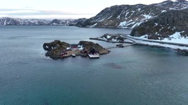 Norveç 'in sakin Arktik ada köyünde, kıyı boyunca, karla kaplı dağlara ve durgun mavi sulara karşı kurulan, yürüyüş yollarıyla birbirine bağlı kırmızı ahşap kulübeler yer alıyor.