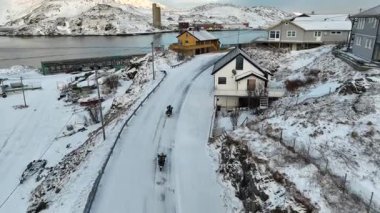 Kavisli bir yolu olan karlı yamaçlı sahil köyü sahnesi rıhtımda tekneler, uzakta su ve renkli evler. Motorsikletler karla kaplı yolda gidiyor. Norveç