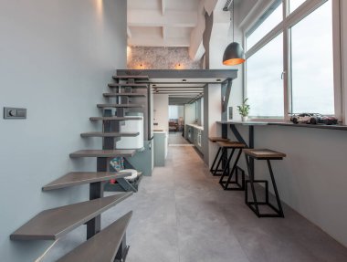 Modern bir apartman dairesi içinde asma kata çıkan şık bir merdiven, minimalist mutfak dolabı ve geniş pencerelerden geçen parlak doğal ışıklandırma var..