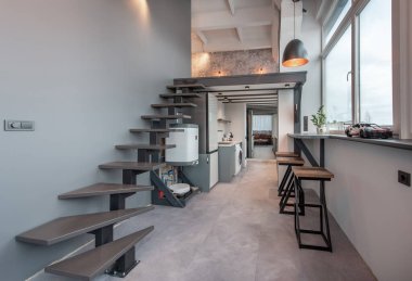 Modern bir apartman dairesi içinde asma kata çıkan şık bir merdiven, minimalist mutfak dolabı ve geniş pencerelerden geçen parlak doğal ışıklandırma var..