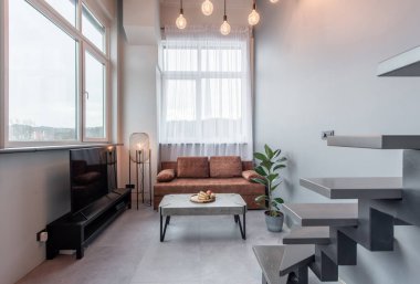 Parlak ve zarif bir oturma odasında minimalist bir dekor, doğal ışık dolu bir ortam ve sanayi tipi detaylar ve modern estetikle zıtlaşmış rahat bir kanepe var.