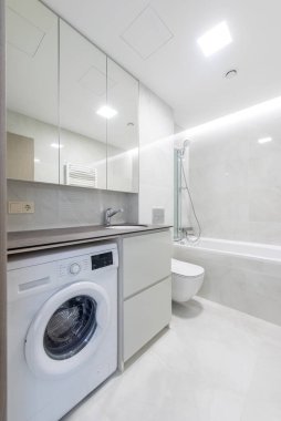 Küvet, çamaşır makinesi ve bol ışıklandırmalı zarif beyaz ve gri dekor unsurları olan parlak ve temiz çağdaş bir banyo. Etkili işlevsellik ve modern estetik için tasarlandı.