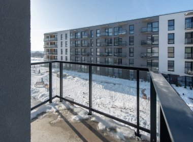 Güneşli bir kış günü öğleden sonra karla kaplı modern bir apartman kompleksinin avlusunu görüntüleyen balkondan kentsel yaşamı ve mevsimsel cazibeyi resmeden bir manzara.