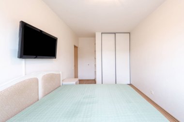 Monte edilmiş televizyon, minimalist mobilyalar ve yumuşak mavi yatak örtüsü içeren temiz ve basit bir yatak odası tasarımı. Modern ya da minimalist iç tasarım kavramları için ideal.