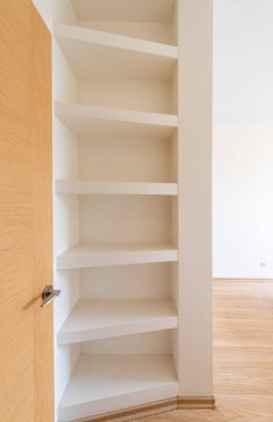 Çağdaş bir yaşam alanının ahşap zeminini tamamlayan, parlak ahşap kapıya bitişik, temiz, beyaz bir raf sistemi gösteren bir köşe ünitesi.