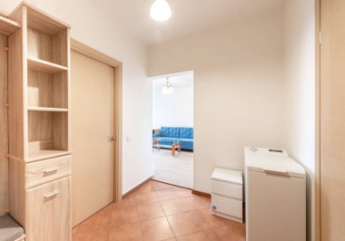 Ahşap kapılar, işlevsel depolama mobilyaları, fayans döşeme ve arka planda yatak odası içeren parlak ve düzenli bir giriş.