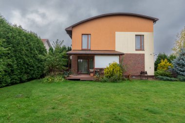 Gri bulutlu gökyüzünün altında canlı yeşil çimenler, çalılar, ağaçlar ve bitişik evler barındıran bir yerleşim yeri..