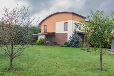 Gri bulutlu gökyüzünün altında canlı yeşil çimenler, çalılar, ağaçlar ve bitişik evler barındıran bir yerleşim yeri..