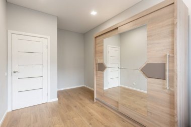 Ahşap döşemeleri olan modern boş bir oda, büyük aynalı bir gardırop, ve pencereden geçen doğal gün ışığı.