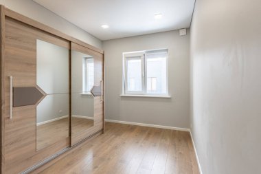 Ahşap döşemeleri olan modern boş bir oda, büyük aynalı bir gardırop, ve pencereden geçen doğal gün ışığı.