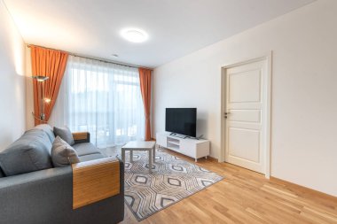 Parlak ve davetkar oturma odası rahat bir kanepe, ahşap zemin, modern mobilyalar ve zarif turuncu perdelerle geniş pencerelerden yayılan doğal ışık.