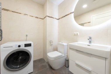 Önden yükleme makinesi, beyaz tuvalet ve modern fayanslı duvarları olan parlak ışıklı bir banyo. Sıkışık iç mekanlarda kullanım alanı ve biçimi birleştirmede etkin