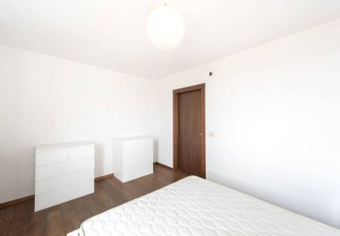 Beyaz mobilyalar, ahşap döşemeler ve basitliği ve modern tasarım unsurlarını vurgulayan düzensiz bir dekorun yer aldığı parlak ve minimalist yatak odası.