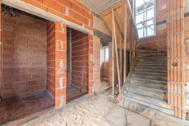 Tamamlanmamış bir evin içinde kırmızı tuğla duvarlar, açık kablolar ve inşaatta bir merdiven var. İlerleme ve çalışmayı temsil eden görünür mimari elementlere sahip parlak bir çalışma alanı