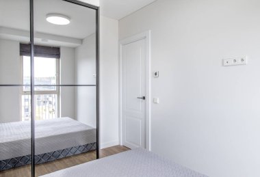 Aynalı dolap kapıları, doğal ışık ve temiz beyaz duvarları olan parlak minimalist yatak odası.