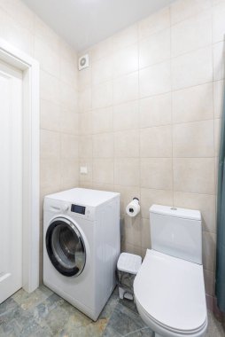 Tuvaletin yanında çamaşır makinesi olan parlak ve temiz bir banyo. Oda açık bej kaplama duvarlar içeriyor, çağdaş bir tasarım.