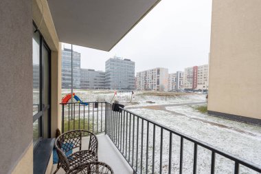 Karlı bir kış gününde modern apartman binalarına bakan rahat bir balkon, dinlenmek için ayarlanmış sandalyeler ve sakin bir açık hava ortamı.