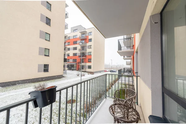 Karlı bir kış gününde modern apartman binalarına bakan rahat bir balkon, dinlenmek için ayarlanmış sandalyeler ve sakin bir açık hava ortamı.