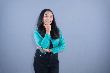 Deniz mavisi ceketli gülümseyen genç bir kadın eli çenesinde gri bir arkaplanda poz veriyor.