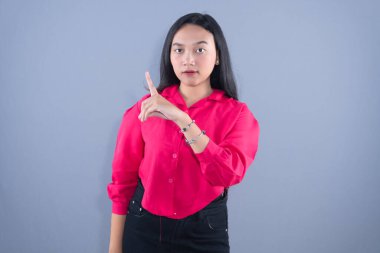 Pembe gömlekli genç bir kadın parmağını gri arka planda gösteriyor.