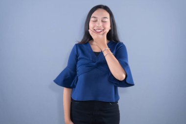 Mavi bluzlu genç bir kadın eli çenesinde gri bir arka planda gülümsüyor.