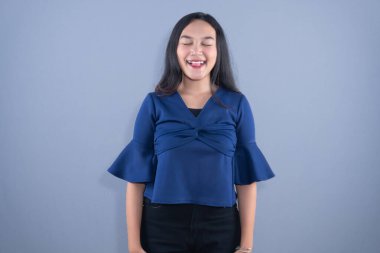 Mavi bluzlu genç bir kadın gülümsüyor, gözleri kapalı, dilini dışarı çıkarıyor.