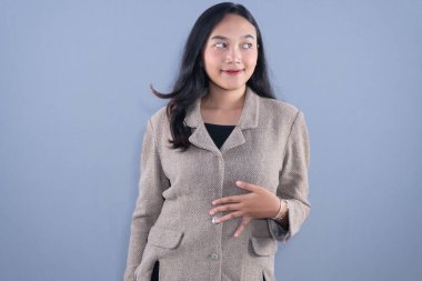 Bej ceketli genç bir kadın karnını tutarken yan bakıyor ve gülümsüyor.