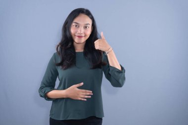 Koyu yeşil bluzlu, gülümseyen bir kadın başparmağını kaldırır ve karnını tutar.