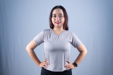 Gri tişörtlü gülümseyen bir kadın elleri kalçasında durur.