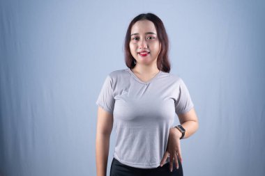 Gri tişörtlü ve siyah popolu gülümseyen bir kadın açık bir arkaplanda duruyor.