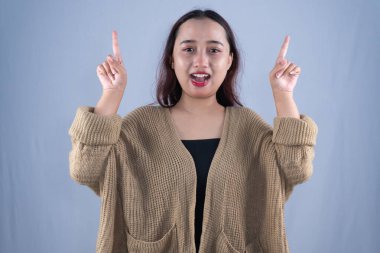 Bej rengi hırkalı, gülümseyen bir kadın iki elini de gri arka planda yukarı kaldırır.