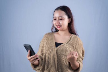 Bej bir hırka giymiş gülümseyen bir kadın elinde bir telefon tutuyor ve bir kutlama hareketi yapıyor.