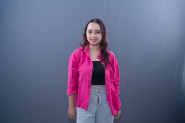 Pembe gömlekli gülümseyen genç bir kadın gri bir arka planda duruyor.