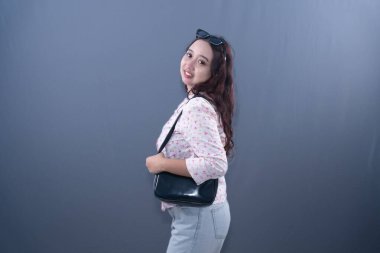 Çiçekli bluzlu ve kot pantolonlu gülümseyen bir kadın iki eliyle siyah bir omuz çantası tutuyor.