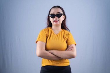 Sarı gömlekli ve kedi gözlü güneş gözlüklü bir kadın kollarını kavuşturarak gülümsüyor.