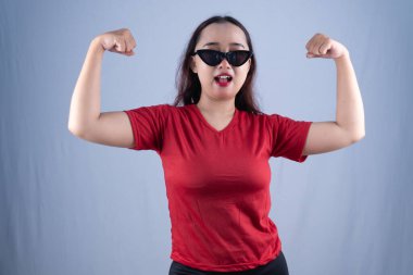 Kırmızı tişörtlü ve siyah kedi gözlü güneş gözlüklü bir kadın kaslarını esnetiyor.