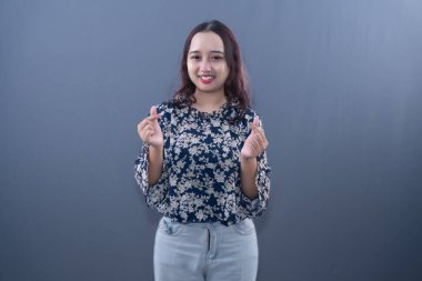 Lacivert çiçekli gömlekli ve kot pantolonlu bir kadın parmak kalp hareketleri yaparken gülümsüyor.