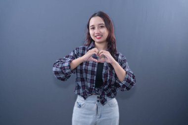 Ekose gömlekli ve kot pantolonlu gülümseyen bir kadın elleriyle kalp işareti yapar.