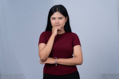 Kestane rengi tişörtlü genç bir kadın eli çenesinde ve ciddi bir ifadeyle ayakta duruyor.