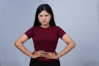 Kestane rengi tişörtlü genç bir kadın elleri kalçasında ve sert bir ifadeyle ayakta duruyor.