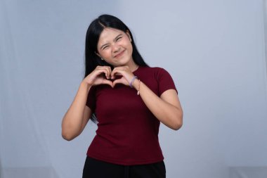 Kestane rengi tişörtlü genç bir kadın elleriyle kalp şekli yapıyor ve gülümsüyor.